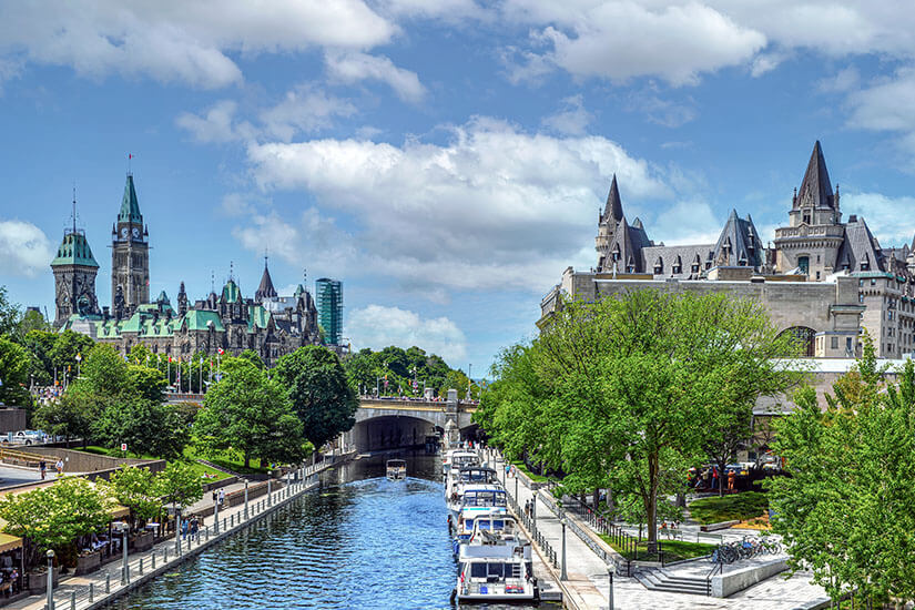 Ein Kanal fließt durch eine Stadt. Rechts liegen Boote am Ufer, im Hintergrund sind Gebäude mit Türmen und eine Brücke zu sehen. Kanada Ottawa