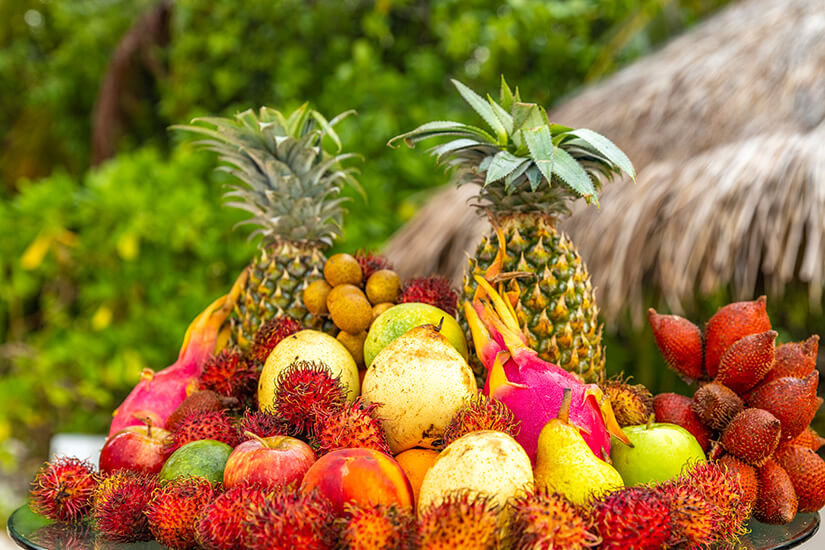 Eine üppige Auslage exotischer und heimischer Früchte ist vor einer tropischen Vegetation und einem strohgedeckten Dach arrangiert. Malediven Essen Obst