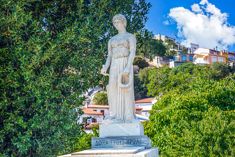 Eine weiße Marmorstatue einer Frau in antikem Gewand, die einen Kranz in der Hand hält, steht auf einem Sockel mit griechischer Inschrift vor einem dichten grünen Baum, während im Hintergrund die Hänge des Dorfes Agiasos mit hellen Häusern unter blauem Himmel aufragen. Lesbos Griechenland Sappho in Agiasos