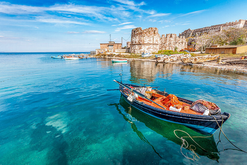 Ein traditionelles Fischerboot mit orangefarbenen Netzen liegt im kristallklaren, türkisfarbenen Wasser, das den Blick bis auf den Meeresgrund freigibt. Im Hintergrund erstreckt sich am flachen Ufer die historische Festung von Mytilini mit ihren alten Steinmauern und Wehrtürmen unter einem weiten, leicht bewölkten Himmel. Lesbos Griechenland Mytilini Hafen