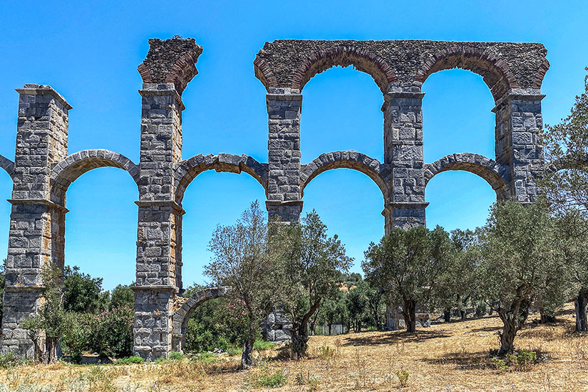Die monumentalen Überreste des römischen Aquädukts von Moria zeigen hohe, mehrstöckige Steinbögen aus grauen Quadern, die teilweise unvollständig in den Himmel ragen. Vor den antiken Ruinen stehen knorrige grüne Olivenbäume auf trockenem, gelblichem Grasland unter einem wolkenlosen, intensiv blauen Himmel. Lesbos Griechenland Aquaedukt von Moria