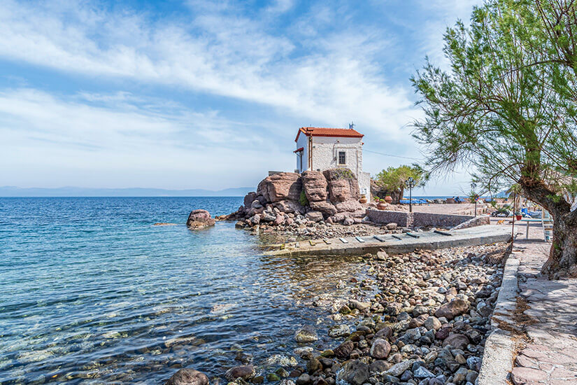 Ein Steinstrand mit bunten Kieselsteinen im Vordergrund führt zu einem klaren, bläulich schimmernden Meer. Am Ende des Strandes steht die kleine, weiße Kapelle Panagia Gorgona mit rotem Ziegeldach auf einem markanten, dunklen Felsen direkt am Wasser, während rechts ein schattenspendender grüner Baum das Motiv einrahmt. Lesbos Griechenland Strand Sikamineas