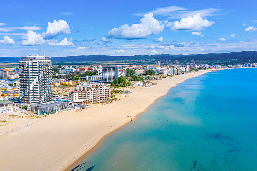 Der Sonnenstrand in Bulgarien ist zu sehen. Heller Sand trifft auf türkisblaues Meerwasser. Hotels und Gebäude reihen sich entlang des Abschnitts. Goldstrand Sonnenstrand Bulgarien Willkommen