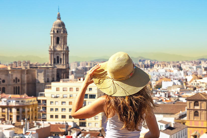 Eine junge Frau mit Strohhut blickt von einem erhöhten Aussichtspunkt auf die sonnendurchflutete Altstadt von Málaga mit dem markanten Glockenturm der Kathedrale und der Bergsilhouette im Hintergrund. Packliste Spanien Frau