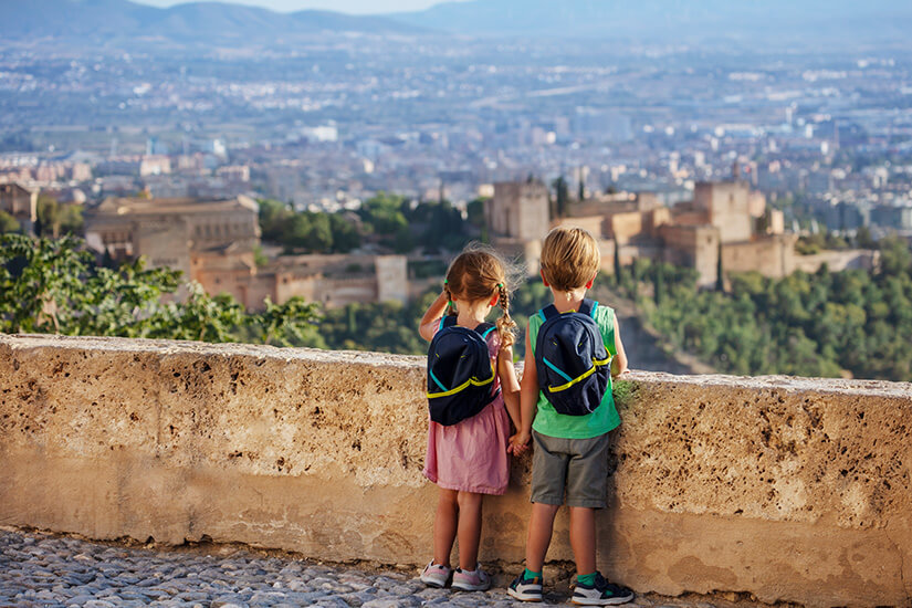 Zwei kleine Kinder mit Rucksäcken stehen Händchen haltend an einer Steinmauer und blicken auf die imposante Alhambra in Granada mit dem Stadtpanorama und der Bergkulisse der Sierra Nevada im Hintergrund. Packliste Spanien Kinder