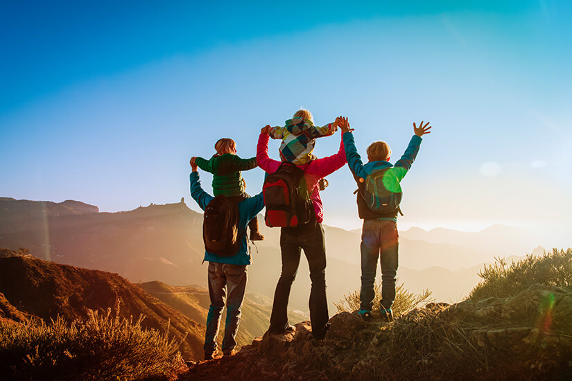 Eine vierköpfige Familie mit Wanderrucksäcken steht in der goldenen Abendsonne auf einem Felsen, die Eltern tragen die jüngeren Kinder auf den Schultern und alle strecken jubelnd die Arme empor vor einer imposanten Berglandschaft. Familie in den Bergen