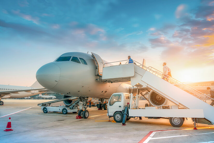 Im warmen Licht der untergehenden Sonne steht ein Passagierflugzeug auf dem Vorfeld bereit, während Fluggäste über eine mobile Gangway einsteigen und das Bodenpersonal letzte Vorbereitungen trifft. Parken am Flughafen Boarding