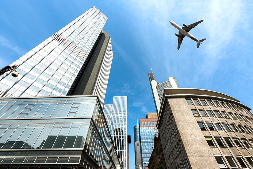 Aus der Froschperspektive ragen die markanten Hochhäuser des Frankfurter Bankenviertels in den blauen Himmel, während ein Passagierflugzeug über die Skyline hinwegfliegt. Parken am Flughafen Frankfurt