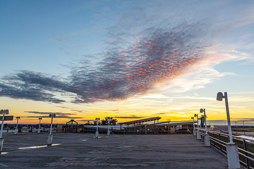Ein leeres Parkdeck mit Straßenlaternen und Geländer öffnet sich zu einem dramatischen Sonnenuntergangshimmel mit rosa-orange leuchtenden Schäfchenwolken über einer flachen Landschaft. Parken am Flughafen Nuernberg