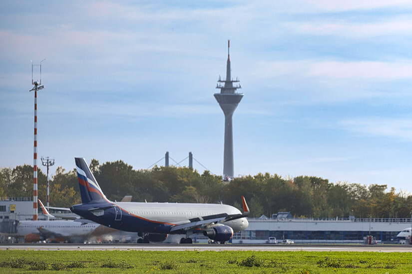 Ein Passagierflugzeug rollt über das Vorfeld eines Flughafens, im Hintergrund ragt der charakteristische Rheinturm in den Himmel. Parken am Flughafen Duesseldorf