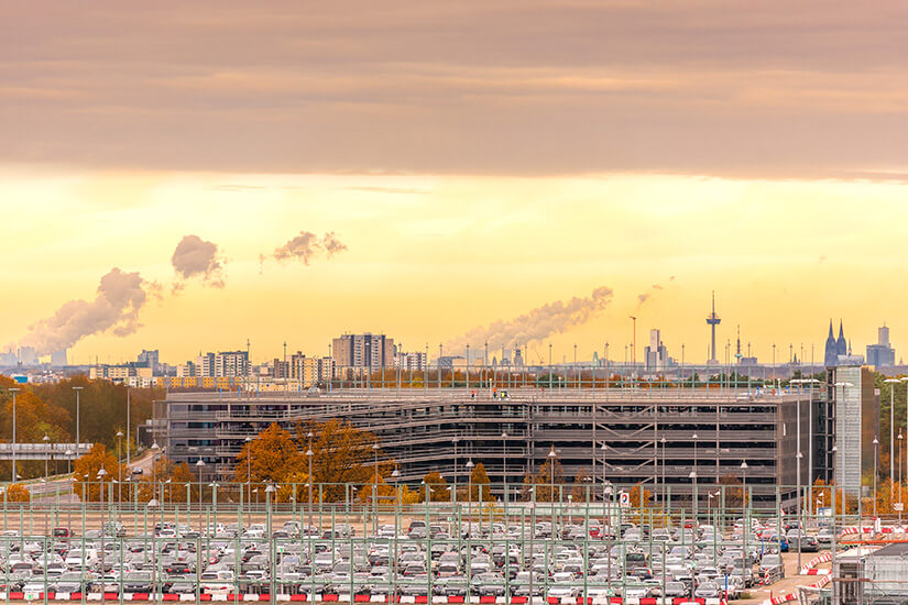 Im warmen Licht eines Herbstabends erstrecken sich Parkplätze mit zahlreichen Fahrzeugen vor einem mehrstöckigen Parkhaus, im Hintergrund zeichnen sich die Silhouette des Kölner Doms, der Colonius-Fernsehturm und qualmende Industrieschornsteine ab. Parken am Flughafen Koeln Bonn