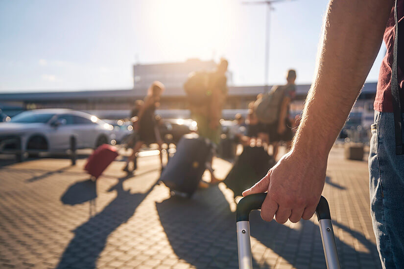 Im Vordergrund umfasst eine Hand den Teleskopgriff eines Trolleys, während im warmen Gegenlicht der Morgensonne weitere Reisende mit ihrem Gepäck Richtung Flughafenterminal eilen. Parken am Flughafen Passagier