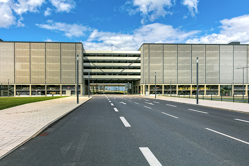 Eine breite, leere Zufahrtsstraße mit Richtungspfeilen führt unter blauem Himmel mit Schäfchenwolken zwischen zwei modernen, mehrstöckigen Parkhäusern mit metallischer Fassade hindurch. Parken am Flughafen Berlin Brandenburg
