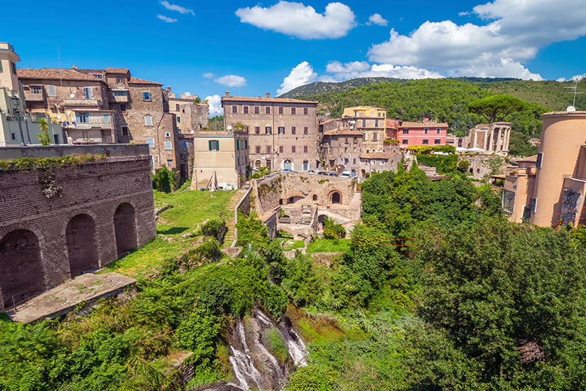 Eine historische Stadtlandschaft mit alten Gebäuden und den Überresten antiker Mauern erstreckt sich über ein grünes Tal. Zwischen den Ruinen fließt ein kleiner Wasserlauf oder Wasserfall durch üppige Vegetation. Im Hintergrund sind bewaldete Hügel und weitere historische Bauwerke zu erkennen. Tivoli Stadt