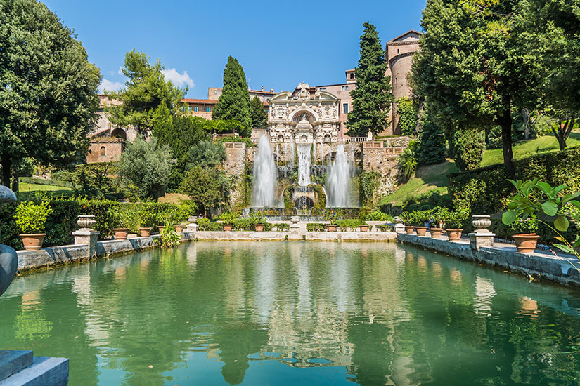 Ein symmetrisch angelegter Renaissancegarten erstreckt sich um ein großes Wasserbecken mit spiegelnder Oberfläche. Im Hintergrund speisen mehrere kunstvolle Brunnen und Wasserspiele eine reich verzierte Fassade, die von üppigem Grün umgeben ist. Tivoli Villa deste