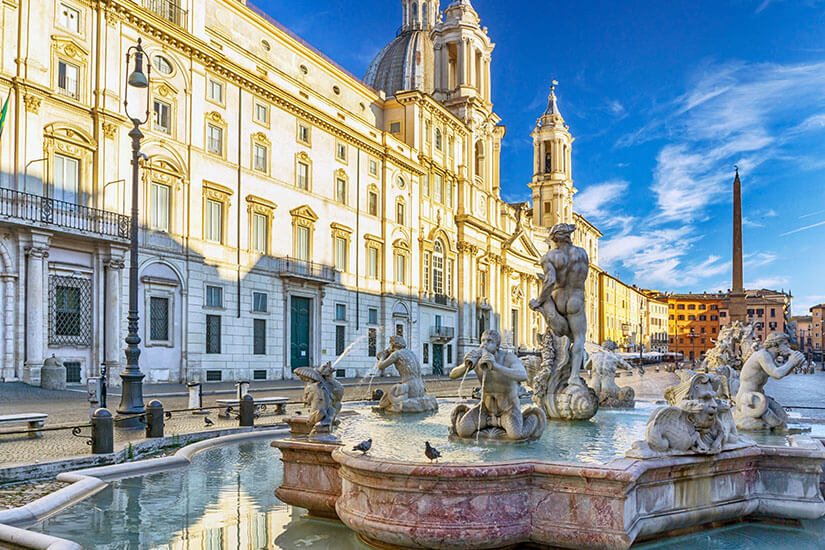 Ein prächtiger Barockplatz wird von einer monumentalen Kirche mit Kuppel und reich verzierter Fassade dominiert. Im Vordergrund befindet sich ein kunstvoll gestalteter Brunnen mit mehreren Marmorstatuen und Wasserspielen. Ein schlanker Obelisk erhebt sich im Hintergrund. Italien Rom