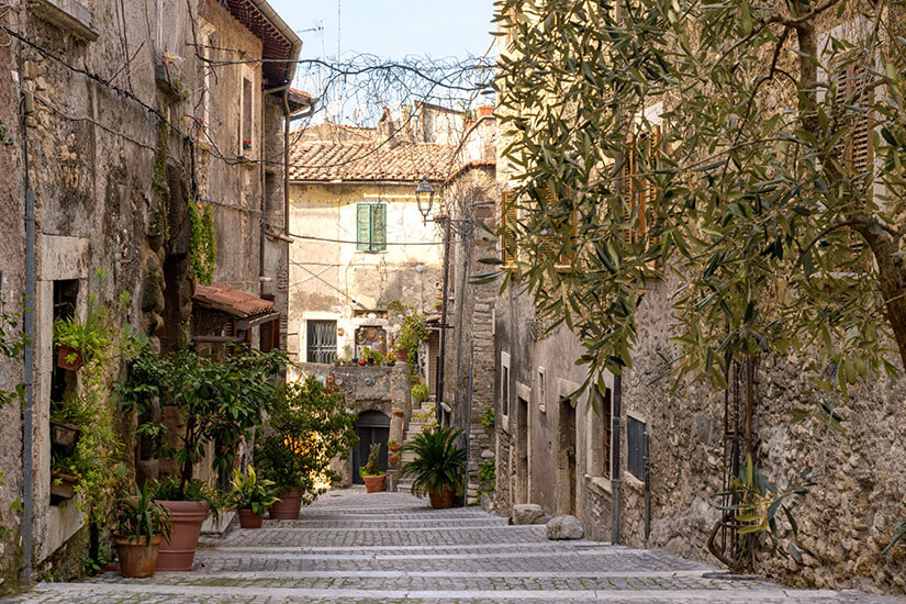 Eine schmale, gepflasterte Gasse führt durch einen historischen Ort mit alten Steinhäusern und verwitterten Fassaden. Zahlreiche Topfpflanzen, kleine Bäume und ein Olivenbaum verleihen der Szene eine mediterrane und gemütliche Atmosphäre. Tivoli Straßen