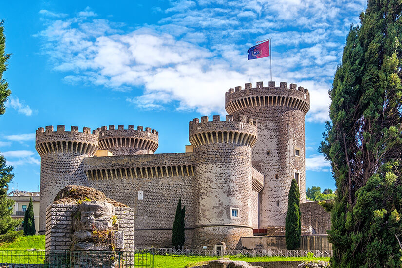 Eine massive mittelalterliche Festung mit mehreren runden Türmen und Zinnen erhebt sich hinter einer Grünfläche. Auf dem höchsten Turm weht eine Flagge. Tivoli Rocca Pia