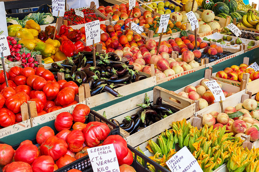 Ein farbenfroher Marktstand bietet eine große Auswahl an frischem Obst und Gemüse in Holzkisten an. Zu erkennen sind unter anderem Tomaten, Auberginen, Paprika, Pfirsiche, Melonen und Zucchiniblüten, jeweils mit handgeschriebenen Preisschildern versehen. Latium Markt