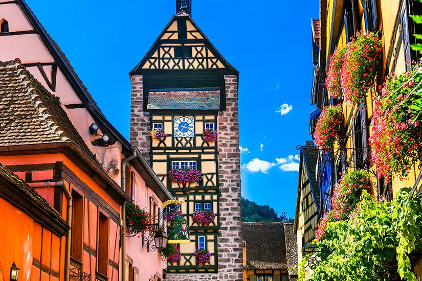 Blick durch eine schmale Gasse auf den historischen Dolderturm in Riquewihr, ein hohes Fachwerktor aus rötlichem Naturstein mit einer großen Uhr und reich geschmückten Blumenkästen an den Fenstern. Riquewihr Frankreich Dolder