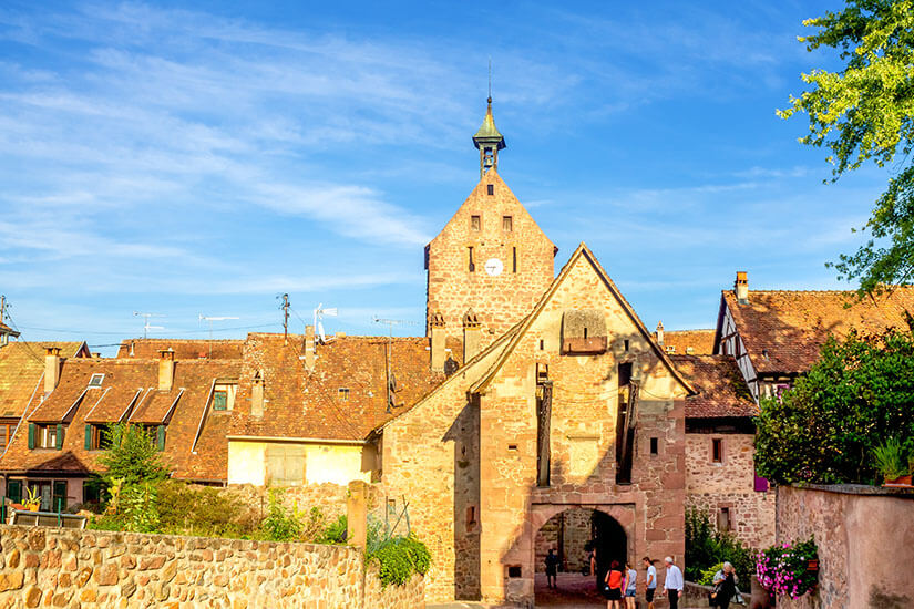 Das historische Obertor von Riquewihr aus rötlichem Sandstein präsentiert sich mit einem großen, offenen Torbogen, durch den Passanten gehen, und einer hölzernen Zugbrückenkonstruktion an der Außenseite. Riquewihr Frankreich Obertor