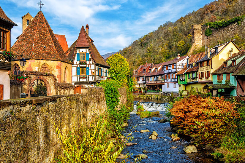 Ein fließender Fluss mit Steinen und kleinen Stromschnellen wird von einer alten Steinmauer und historischen Fachwerkhäusern in bunten Farben wie Gelb, Grün und Weiß gesäumt. Auf der linken Seite steht ein markantes Fachwerkhaus mit blauen Fensterläden neben einer kleinen Kapelle, während im Hintergrund ein bewaldeter Hang mit den Überresten einer alten Burgmauer unter einem leicht bewölkten Himmel emporragt. Riquewihr Frankreich Kaysersberg
