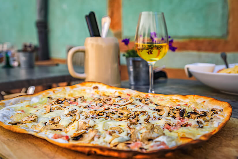 Ein großer, knusprig gebackener Flammkuchen mit dünnem Teigrand, reichlich belegt mit geschnittenen Champignons, Zwiebeln und Speck, liegt auf einem Holzbrett auf einem Tisch im Freien. Im unscharfen Hintergrund stehen ein gefülltes Glas elsässischer Weißwein, ein Steinkrug mit Besteck und ein kleiner Metalleimer mit violetten Blumen vor einer hellgrünen Fachwerkhauswand. Riquewihr Frankreich Flammkuchen