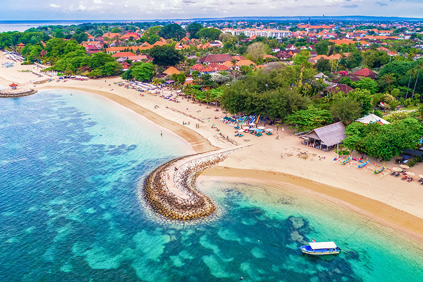 Die geschwungene Sandbucht Sanur auf Bali mit türkisklarem Wasser und einem charakteristischen Steinmolo. Am Strand reihen sich Sonnenschirme und kleine Hütten auf, dahinter erstreckt sich eine dicht bebaute Ortschaft mit typisch balinesischen Dächern. Indonesien Inseln Bali