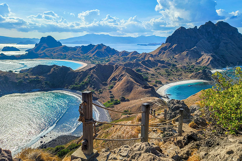 Vom Gipfel der Padar-Insel eröffnet sich ein Blick auf drei Buchten mit unterschiedlich gefärbtem Wasser, eingerahmt von trockenen braunen Hügeln. Ein hölzerner Aussichtspunkt mit Seilgeländer markiert das Ende des Aufstiegspfades. Padar gehört zum Komodo-Nationalpark und gilt als eines der ikonischsten Panoramen Indonesiens. Indonesien Inseln Padar