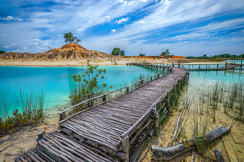 Ein alter Holzsteg führt über türkisblaues Wasser entlang einer durch Bergbau geprägten Landschaft mit weißen Sedimentwänden. Die Szenerie wirkt verlassen, während die Natur das frühere Industriegelände langsam zurückerobert. Das warme Licht verleiht dem Bild trotz seiner Rohheit eine eigene Schönheit. Indonesien Inseln Mangroven