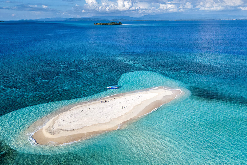 Eine winzige Sandbank liegt nahezu allein im strahlend blauen Meer der Nordmolukken. Das flache, ovale Eiland ist kaum bewachsen und wird von türkis-blauem Wasser umspült, das die Korallenriffe durchscheinen lässt. Ein einzelnes Boot am Rand der Sandbank unterstreicht die Abgelegenheit dieses Ortes. Indonesien Inseln Dodola