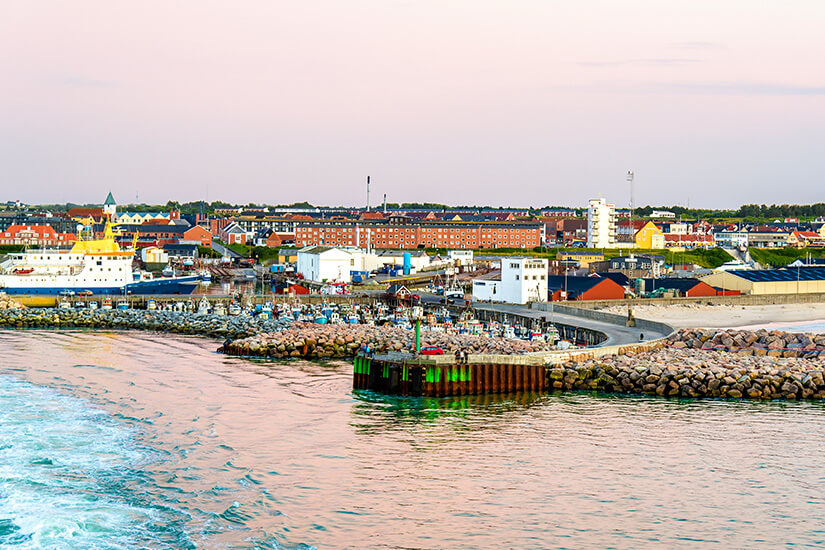Ein Küstenhafen mit Kaianlagen, Wellenbrechern und mehreren Schiffen erstreckt sich entlang des Ufers. Dahinter liegen Wohn- und Industriegebäude, die sich bis zum Horizont ausbreiten. Hirtshals Daenemark Stadt
