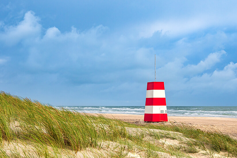 Ein rot-weiß gestreifter See- oder Strandmarkierungsturm steht auf einem sandigen Küstenabschnitt mit Dünengras. Im Hintergrund sind das Meer mit Wellen und ein wolkenverhangener Himmel zu sehen. Hirtshals Daenemark Tornby Strand