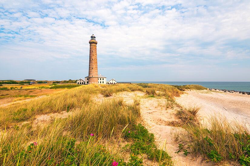 in hoher Leuchtturm erhebt sich über einer Küstenlandschaft mit Sanddünen und trockenem Gras. In seiner Nähe stehen einige Gebäude, während sich rechts das Meer bis zum Horizont erstreckt. Juetland Skagen