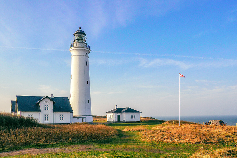 Ein weißer Leuchtturm steht auf einer grasbewachsenen Küstenlandschaft und wird von mehreren Gebäuden flankiert. Daneben weht eine Flagge auf einem hohen Mast, während im Hintergrund das Meer zu sehen ist. Hirtshals Daenemark Leuchtturm