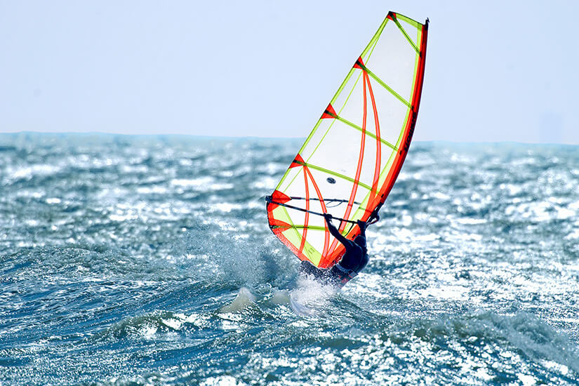 Eine Person fährt auf einem Windsurfbrett über die bewegte Meeresoberfläche und nutzt ein auffällig buntes Segel. Spritzendes Wasser und die geneigte Haltung deuten auf kräftigen Wind und hohe Geschwindigkeit hin. Juetland Surfen