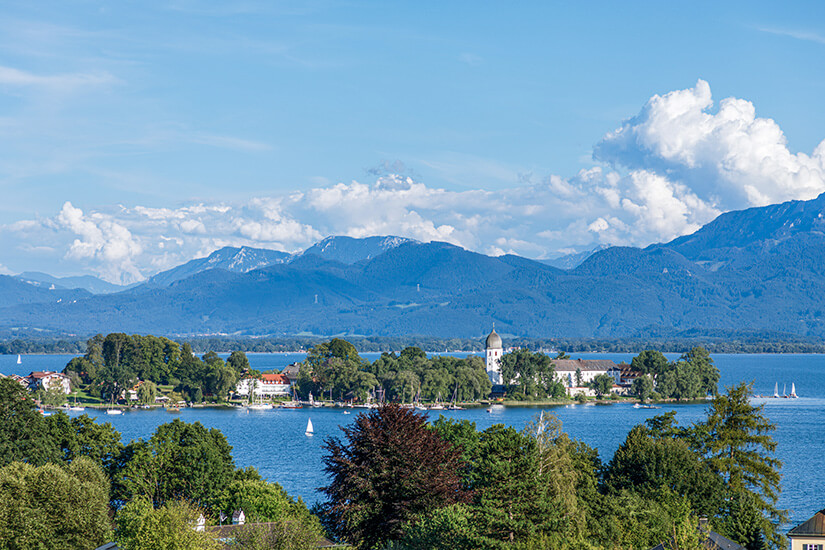 Blick über den Chiemsee auf die Fraueninsel mit dem charakteristischen Glockenturm des Klosters Frauenwörth. Im Hintergrund erheben sich die Chiemgauer Alpen, davor liegt die kleinere, bewaldete Krautinsel. Die Aufnahme zeigt die typische Postkartenkulisse des bayerischen Voralpenlandes. Chiemsee Deutschland Fraueninsel
