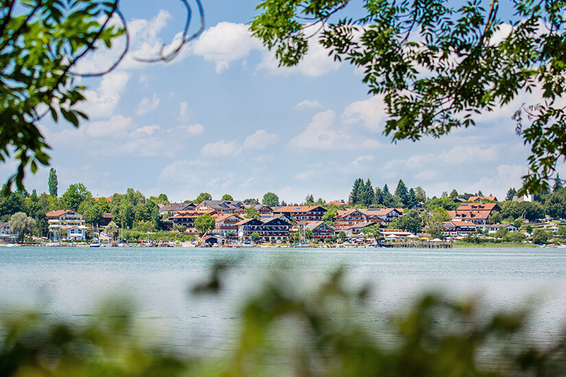 Blick über den See auf die Uferpromenade des Ortes Gstadt am Chiemsee. Die typisch bayerischen Häuser mit roten Ziegeldächern reihen sich entlang des Wassers. Von hier aus starten Schiffe zur gegenüberliegenden Fraueninsel. Chiemsee Deutschland Gstadt