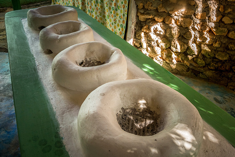In einer Reihe stehen vier traditionelle Lehmöfen mit runden Öffnungen und Asche im Inneren auf einer grün gestrichenen Holzbank vor einer rustikalen Natursteinmauer. Dominikanische Republik Essen Lehmofen