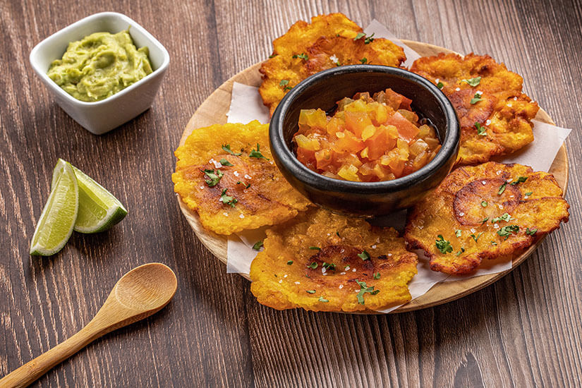 Auf einem Holzteller sind goldgelb frittierte und plattgedrückte Kochbananenscheiben angerichtet, dazu eine schwarze Schale mit Tomaten-Zwiebel-Salsa, eine kleine Schüssel Guacamole und Limettenspalten. Dominikanische Republik Essen Tostones