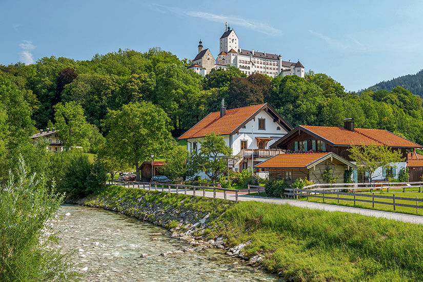 Schloss Hohenaschau thront hoch über dem Ort Aschau im Chiemgau auf einem bewaldeten Felsen. Im Vordergrund fließt die Prien vorbei an typischen bayerischen Häusern mit Holzbalkonen. Die Burg gehört zu den ältesten und größten Höhenburgen Bayerns. Chiemsee Deutschland Schloss