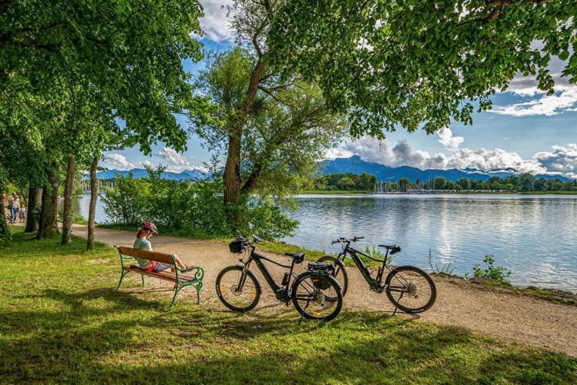 Eine ruhige Rastmöglichkeit am Ufer des Chiemsees mit zwei E-Bikes und einer Bank im Schatten alter Bäume. Im Hintergrund sind das Alpenpanorama und Segelboote zu erkennen. Der Uferweg eignet sich ideal für Radtouren rund um den See. Chiemsee Deutschland Feldwies