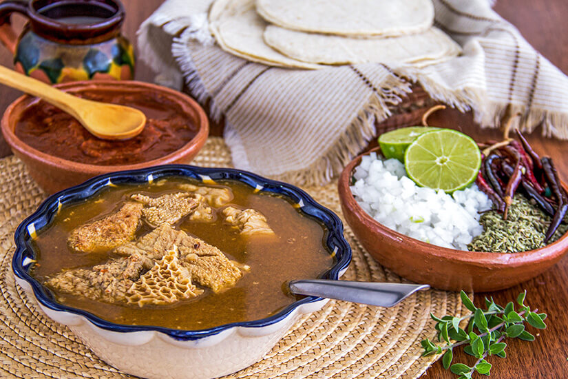 In einer blau verzierten Keramikschale dampft eine kräftige bräunliche Brühe mit Kuttelstücken (Mondongo), serviert mit gehackten Zwiebeln, Limetten, getrockneten Chilis, Oregano in einer Tonschale sowie Tortillas und einer Schale Salsa. Dominikanische Republik Essen Mondongo