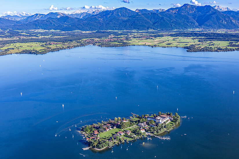 Der Chiemsee mit der Fraueninsel im Vordergrund und dem Alpenpanorama am Horizont. Zahlreiche Segelboote sind auf dem größten See Bayerns unterwegs. Im Hintergrund erstrecken sich die typischen Wiesen und Dörfer des Chiemgaus. Chiemsee Deutschland See