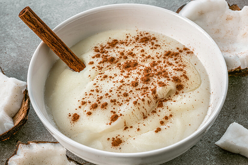 In einer weißen Schale ist eine cremige, weiße Pudding-artige Süßspeise serviert, großzügig mit Zimtpulver bestäubt und mit einer Zimtstange dekoriert, drumherum liegen frische Kokosnussstücke. Dominikanische Republik Essen Majarete