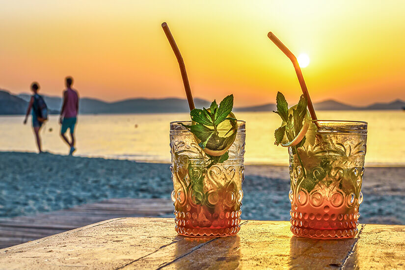 Zwei kunstvoll verzierte Gläser mit Mojito-Cocktails, frischen Minzeblättern, Limettenscheiben und Strohhalmen stehen auf einem Holztisch am Strand, während im Hintergrund ein Paar bei goldenem Sonnenuntergang am Meer entlangspaziert. Dominikanische Republik Essen Getraenke
