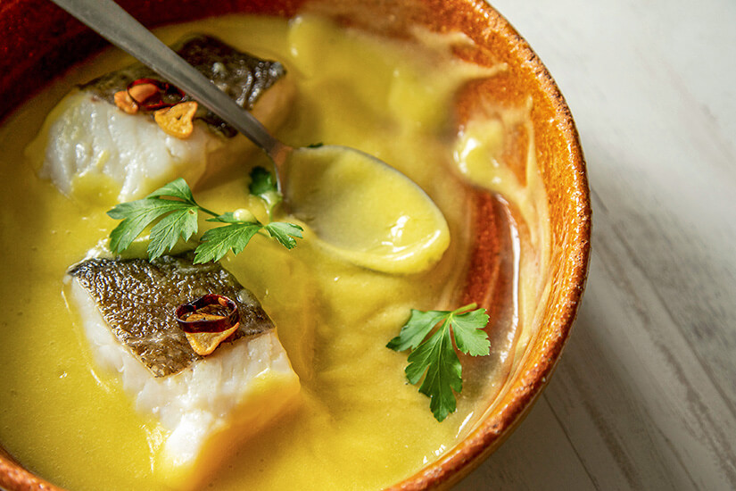 In einer rustikalen Tonschale schwimmen zarte Fischfilets in einer leuchtend gelben Kokos-Curry-Sauce, garniert mit frischer Petersilie und Chili-Knoblauch-Scheiben. Pescado con Coco