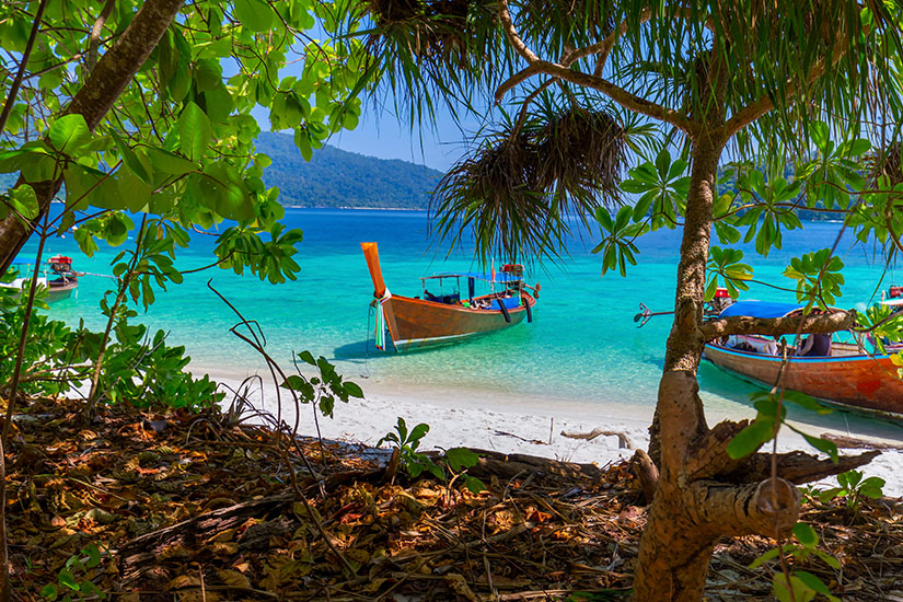 Blick aus dem Schatten dichter, grüner Dschungelbäume und Palmen auf einen feinen, weißen Sandstrand und das glasklare, türkisfarbene Meer. Im seichten Wasser liegen traditionelle thailändische Longtailboote aus Holz vor Anker, während im Hintergrund eine bergige, bewaldete Insel unter einem strahlend blauen Himmel aufragt. Koh Lipe Thailand Willkommen