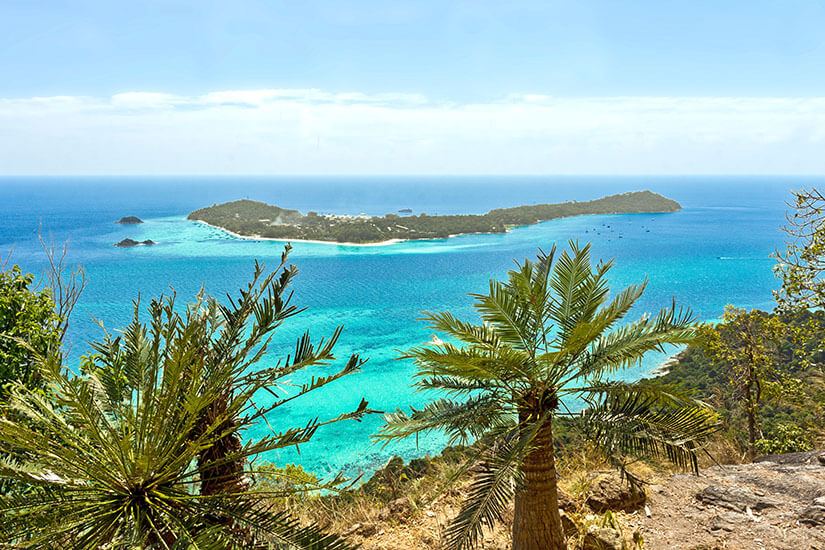 Ein weiter Panoramaausblick von einem hohen, felsigen Aussichtspunkt mit Palmen und tropischen Pflanzen im Vordergrund hinab auf das tiefblaue und leuchtend türkisfarbene Meer. Inmitten des Wassers zeichnet sich die gesamte, flache Nachbarinsel Koh Lipe mit ihren weißen Sandstränden und der grünen Vegetation ab, während am Horizont ein blasser Himmel mit leichten Wolkenstreifen zu sehen ist. Koh Lipe Thailand Ko Adang