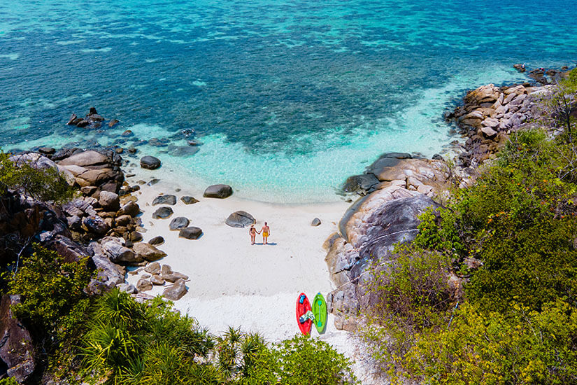 Eine Luftaufnahme einer kleinen, versteckten Sandbucht, die von großen, grauen Felsen und dichtem grünen Gebüsch umschlossen ist. Ein Paar steht Hand in Hand auf dem feinen, weißen Sandstrand und blickt auf das glasklare, flache Meerwasser, das in Türkistönen schimmert, während im Vordergrund ein rotes und ein grünes Kajak am Strand bereitliegen. Koh Lipe Thailand Kajak